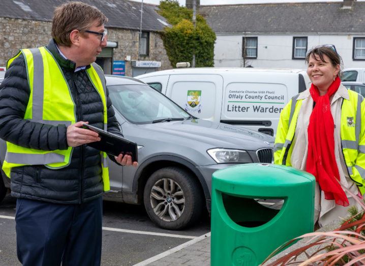 Two people in high-vis jackets are standing beside a bin on a town street.