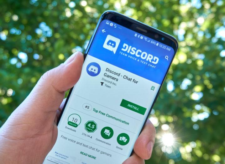 A hand holding a smartphone up against a backdrop of leafy trees. The phone screen has the Discord app on it.