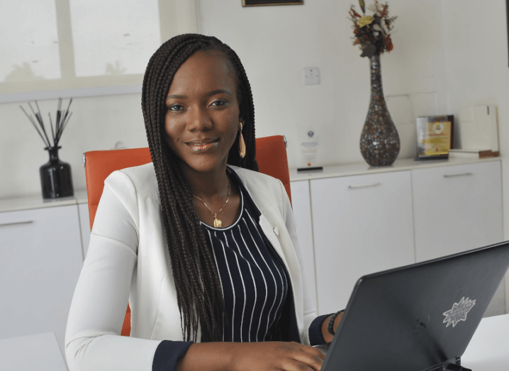 Foyin Olajide-Bello is sitting at a desk with a laptop while smiling at the camera. She’s wearing a white blazer.