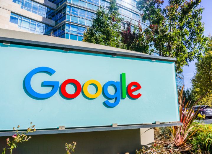 A close-up of the Google logo on a bright blue sign in front of an office building against a blue sky.