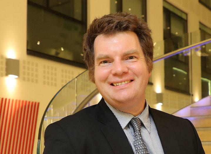 Prof Niall English smiles for a photo next to a staircase. He is wearing a suit and tie.