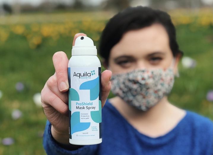 A researcher at Aquila Bioscience is standing outdoors wearing a face mask and holding the ProShield spray up to the camera.