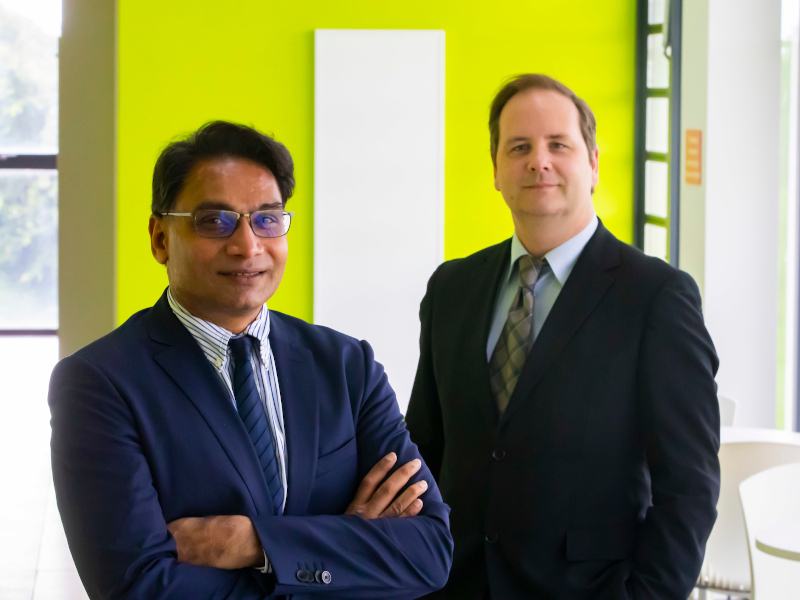 Two men in suits stand together in front of a neon green wall.