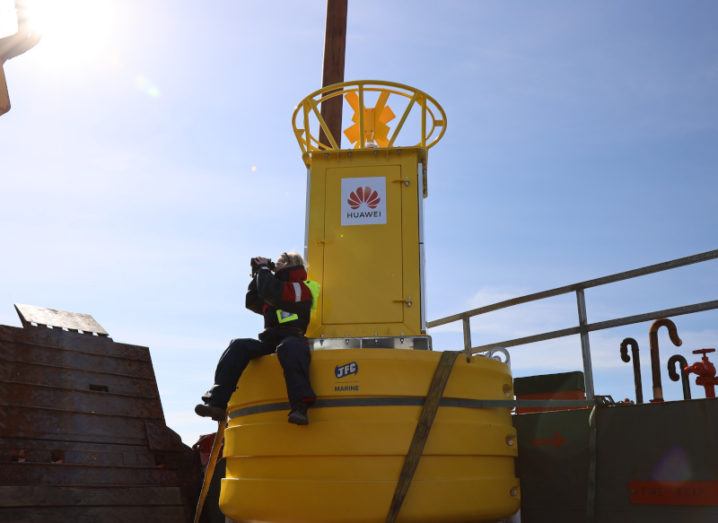 A woman is sitting on a large buoy with Huawei branding while looking through binoculars as part of the Smart Whale Sounds project.