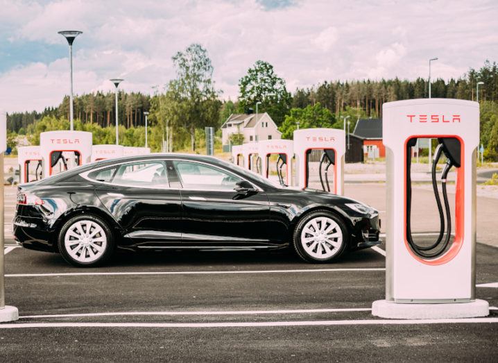 Black Tesla car in an empty car park on a sunny day. The car is surrounded by Tesla-branded charging stations.