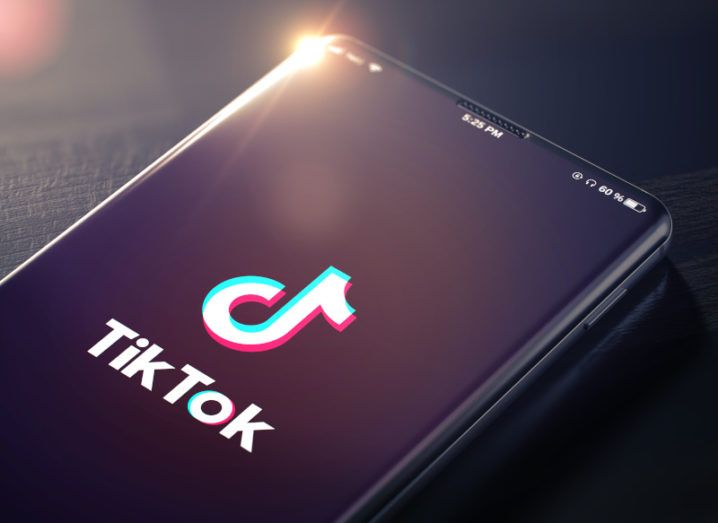 A close-up of a black smartphone on a dark wooden surface. The screen is displaying the TikTok logo.
