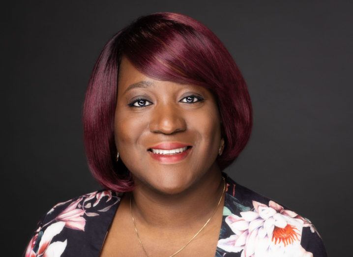 A headshot of Toju Duke smiling at the camera against a black background.