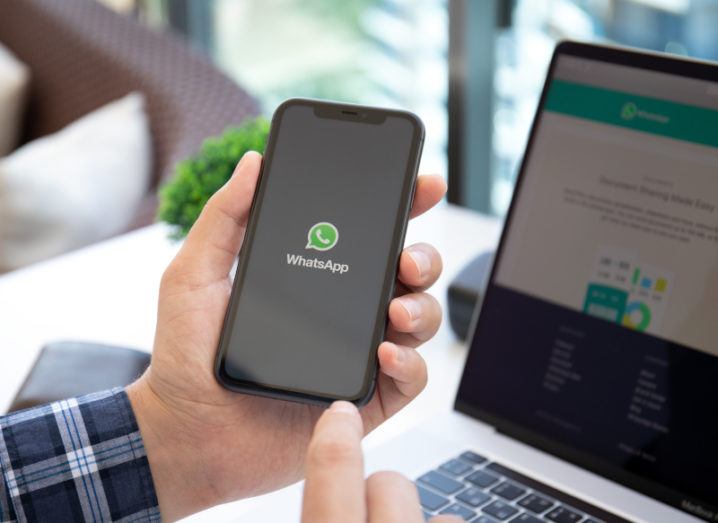 A person holds a smartphone with the WhatsApp logo on it, while a laptop sits in the background with the WhatsApp desktop version open.