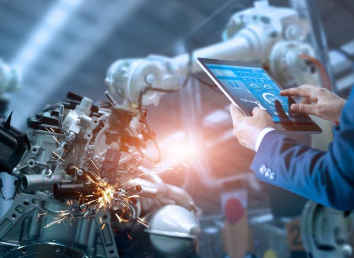 A man in a business suit uses a tablet in front of a large piece of active manufacturing equipment, with sparks flying.