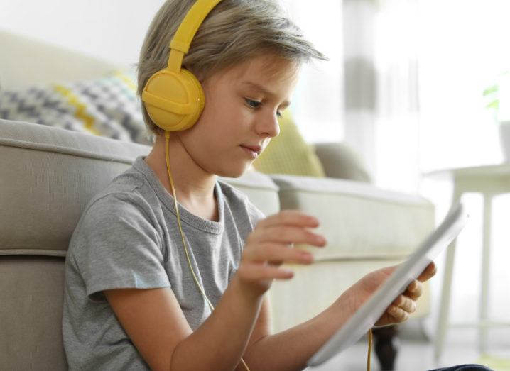 A young blonde boy is wearing yellow headphones while listening to something on a tablet.