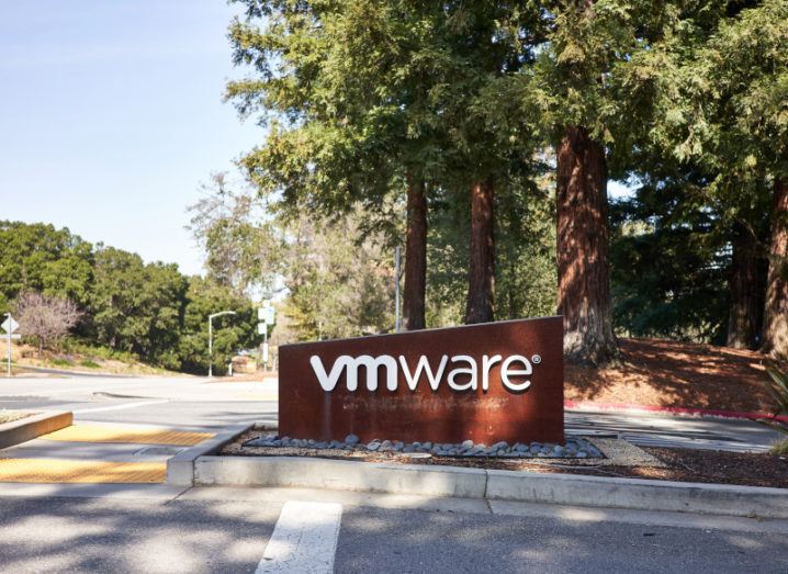 A sign on the side of the road says VMware, beneath the shade of large trees on a sunny day.