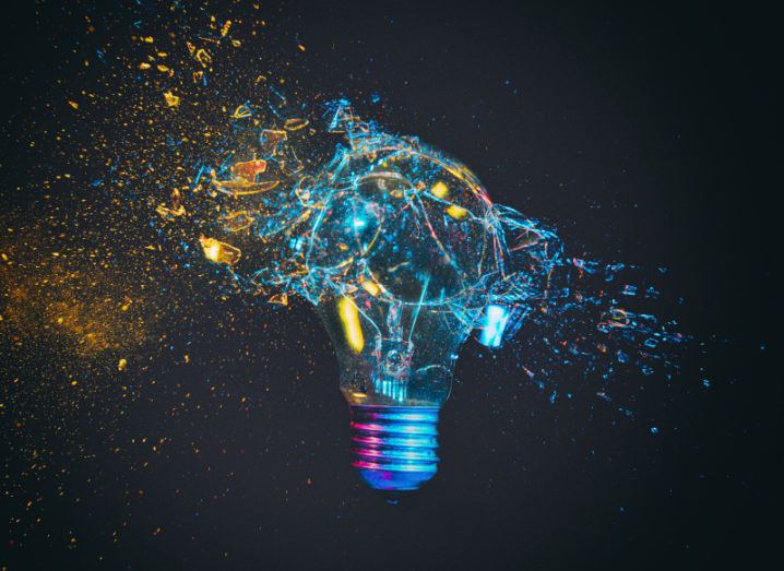 A lightbulb is breaking, throwing shards of glass and flashes of coloured lights against a black background.