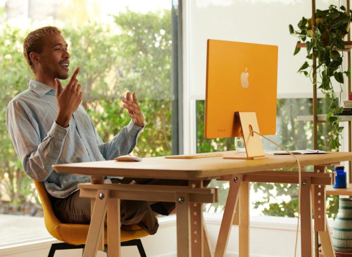 A man sitting at a desk in a brightly lit room while he’s working at a new orange Apple iMac.