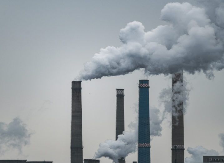 Four industrial chimneys against a dusky grey sky with plumes of smoke coming from them, representing carbon emissions.