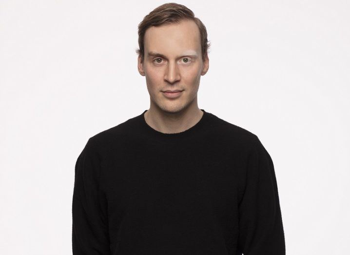 A man in a black sweaters stands against a plain background.