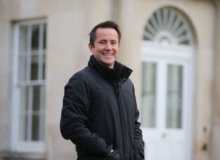 A man stands smiling outside the NovaUCD building on the University College Dublin campus. He has his hands in the pockets of his black coat.