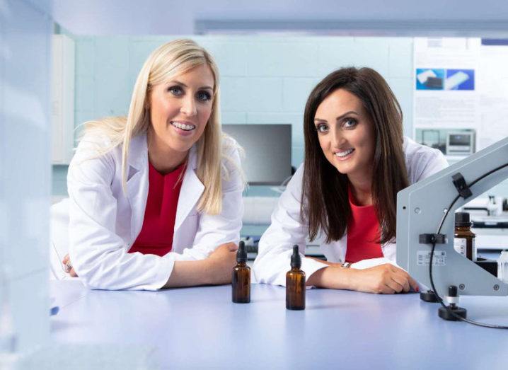 Two women wearing white lab coats are leaning on a countertop in a lab setting.