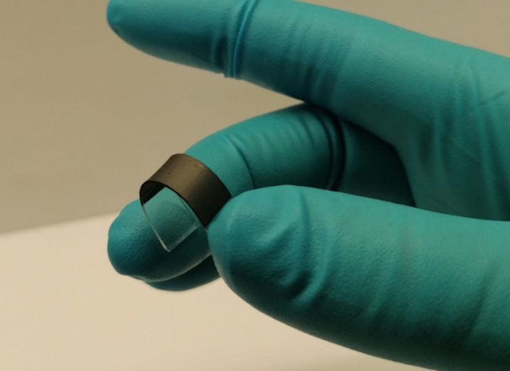 A hand wearing a blue glove is holding a thin flexible material that is coated in a graphene-based ink.