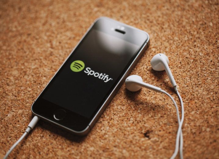 A black smartphone sits on corkboard with white earphones plugged in and lying beside it. The Spotify logo is on the screen.