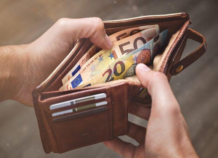 A man’s hands are opening a brown leather wallet, filled with Euro notes.