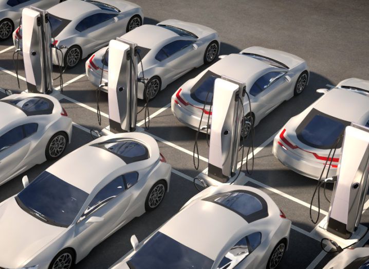 A large aerial view of a car park full of white electric vehicles near electric charging stations.
