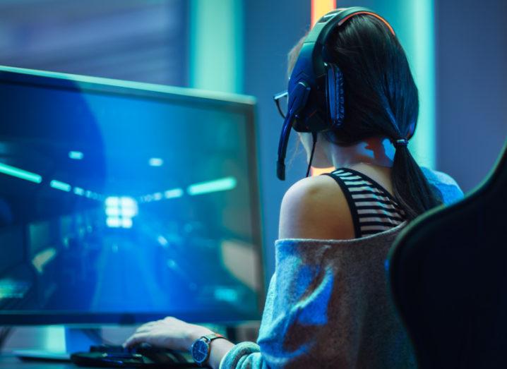 A girl sits in a gaming chair at a large computer screen wearing headphones in a darkened room playing video games.