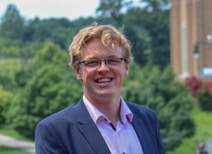 A smiling man stands outside amid some green bushes next to a large building.