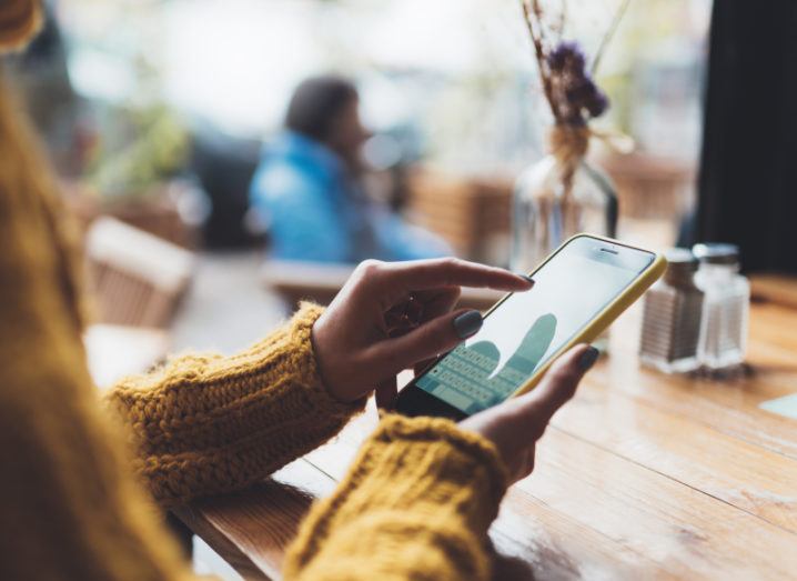 A woman wearing a chunky yellow jumper uses her smartphone in a busy café.