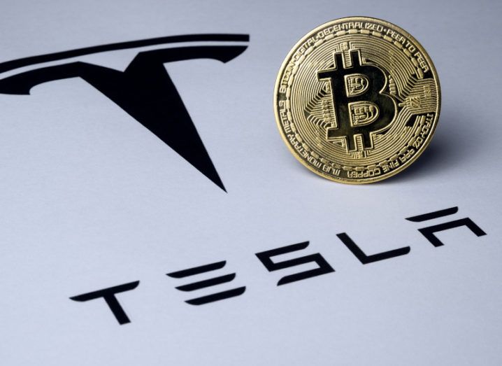 A golden coin bearing the bitcoin logo stands alongside a sheet bearing the Tesla logo.