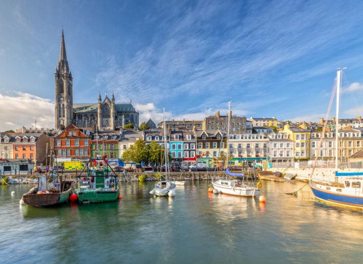 A picturesque photo of Cobh harbour, with several small boats in the water in front of colourful houses and a cathedral, highlighting tourism.