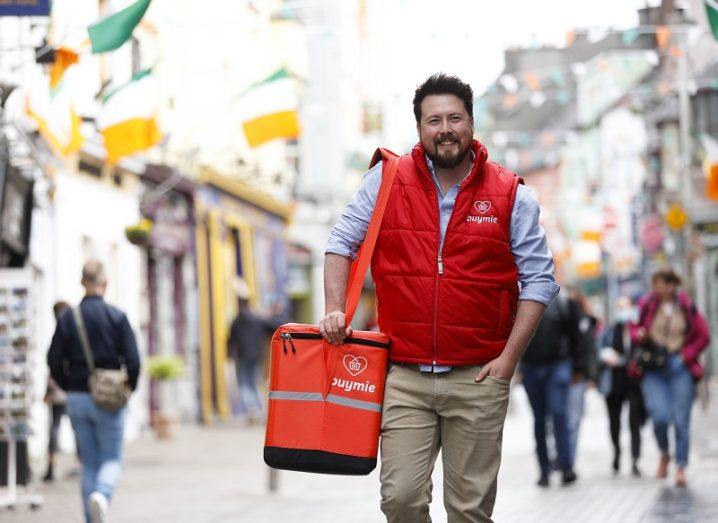 Devan Hughes walks through a city street holding a Buymie delivery bag.
