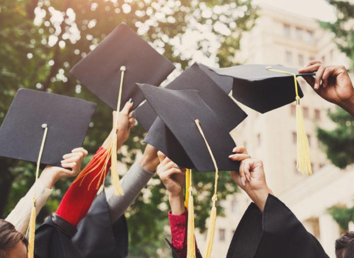 A group of hands holding graduation caps in the air against a backdrop of green trees.