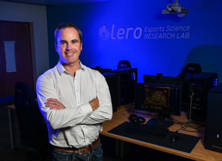 A man in a polkadot shirt stands in a darkened computer lab with Lero Esports Science Research Lab lit up in blue on the back wall.