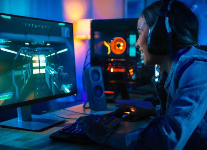 A young woman in a denim jacket plays a video game on a desktop computer with a light-up keyboard and wireless headphones. She is smiling, wholly immersed in the game.