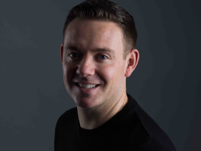A headshot of Dan Harding smiling at the camera against a dark charcoal background.