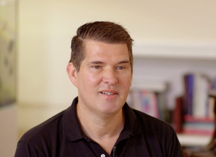 A headshot of a man in a black polo shirt looking slightly off camera while talking.