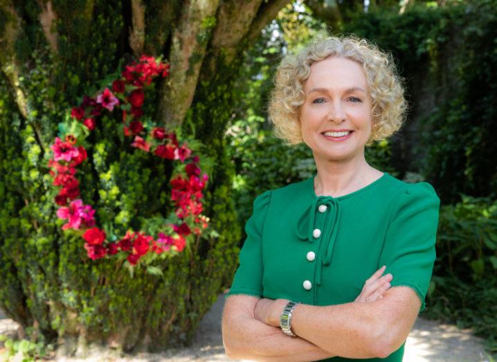 Anne O'Leary stands in front of a tree with red flowers in the shape of the Vodafone logo. She is smiling with her arms crossed and is wearing a green dress.