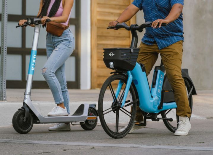 Two people are pictured on the street. A woman is on the left, standing on a Bird brand electric scooter. A man is to her right, seated on a Bird brand electric bike.