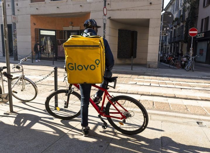 A Glovo delivery rider standing beside his bicycle on the street.