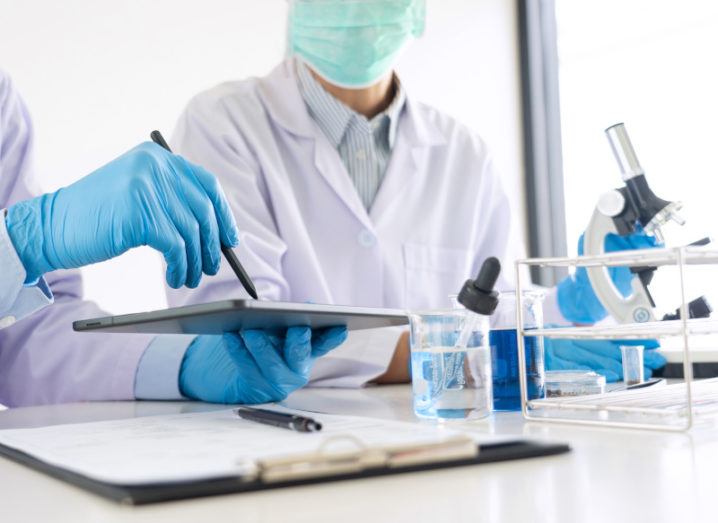 Two scientists sit at a table in lab coats and masks. They are in discussion, and one is holding a pen over a clipboard while the other listens. Research equipment such as glass beakers and a microscope are on the desk in front of them.