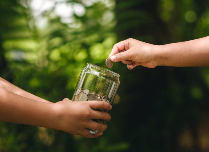 A hand holding a glass jar full of coins out so another hand can place a coin into the jar. The background is a green forest.