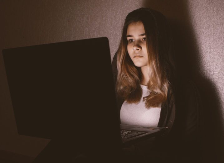 Teenage girl sitting alone in a dark room using a laptop, looking glum.