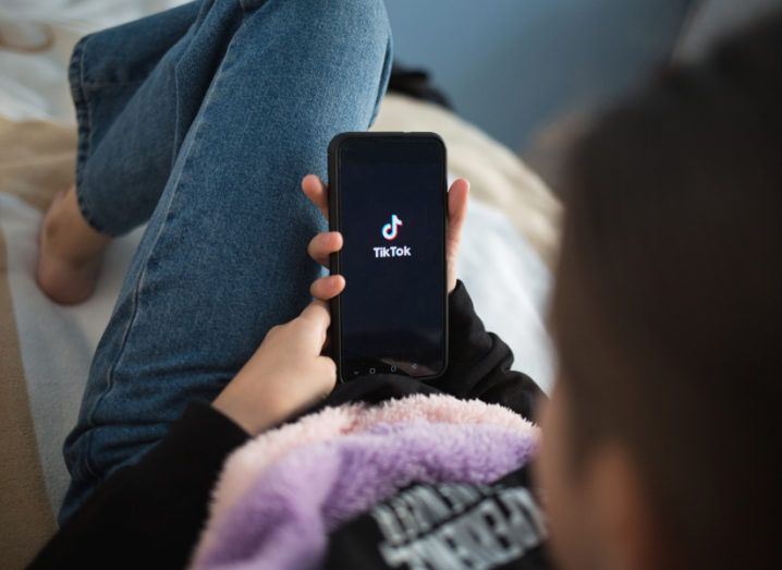 A girl lies on a sofa holding a smartphone in her hand which is displaying the TikTok logo.