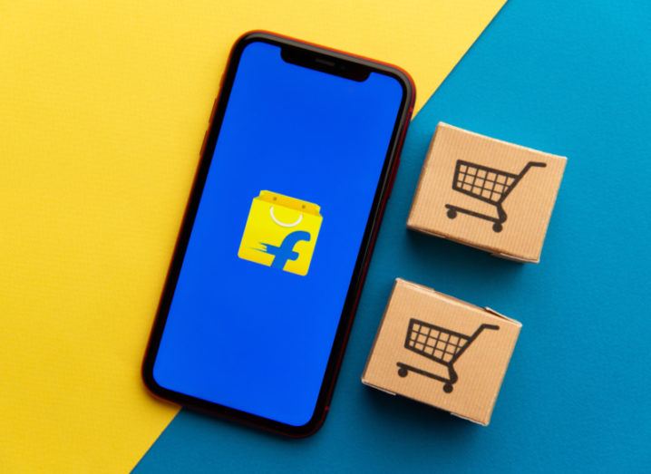 A smartphone displaying the Flipkart logo on its screen lies on a blue and yellow background next to miniature brown boxes marked with shopping cart icons.