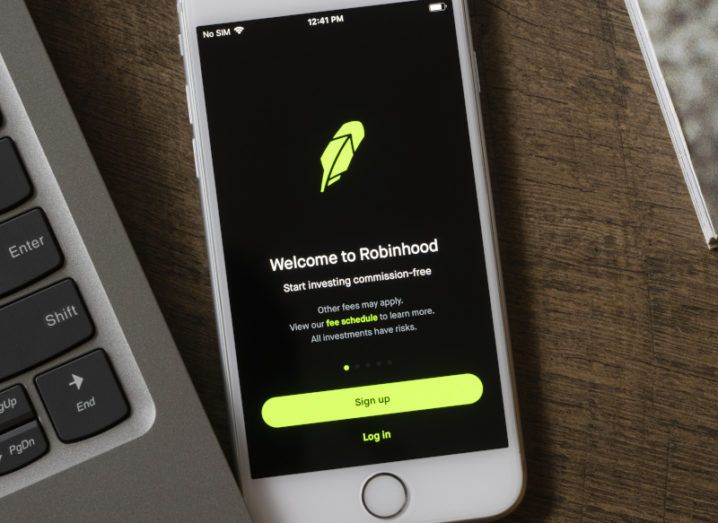 A phone is sitting on a desk beside a laptop. On the screen in the login page for the Robinhood app.