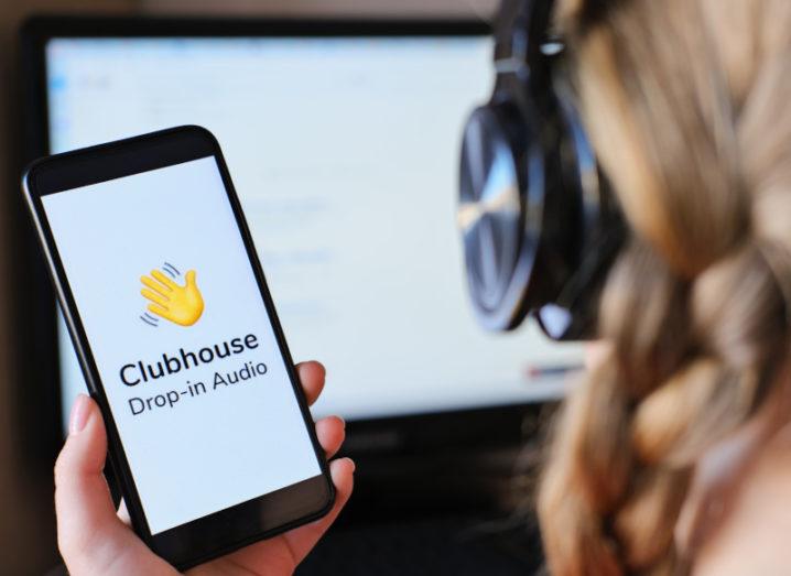 A stock image of a person wearing headphones looking at the Clubhouse app on their phone.