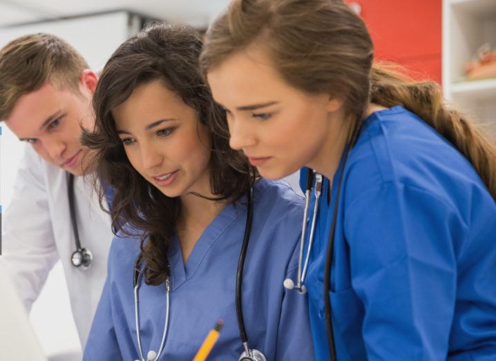 Three young medical doctors gathered together standing around a workstation, which is out of shot. The three young people wear stethoscopes and blue medical scrubs.