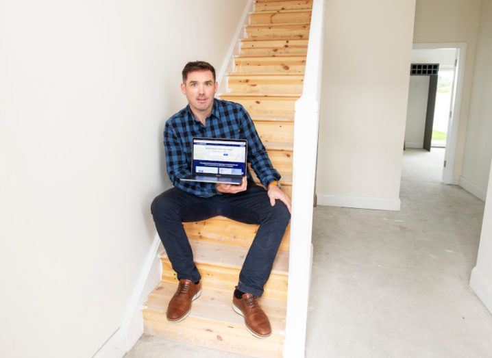 Eoghain Ryan, co-founder of Housebuild.ie, pictured sitting in a stairwell holding a laptop.