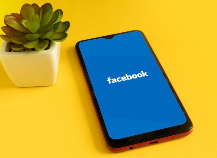 A smartphone lies on a yellow surface beside a small plant. The Facebook logo is on the screen.