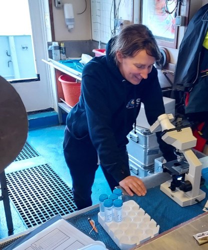 A woman in a tracksuit leans into a lab bench on-board a research vessel, looking at an AI-equipped microscope with a modified iPod Touch as a display screen.